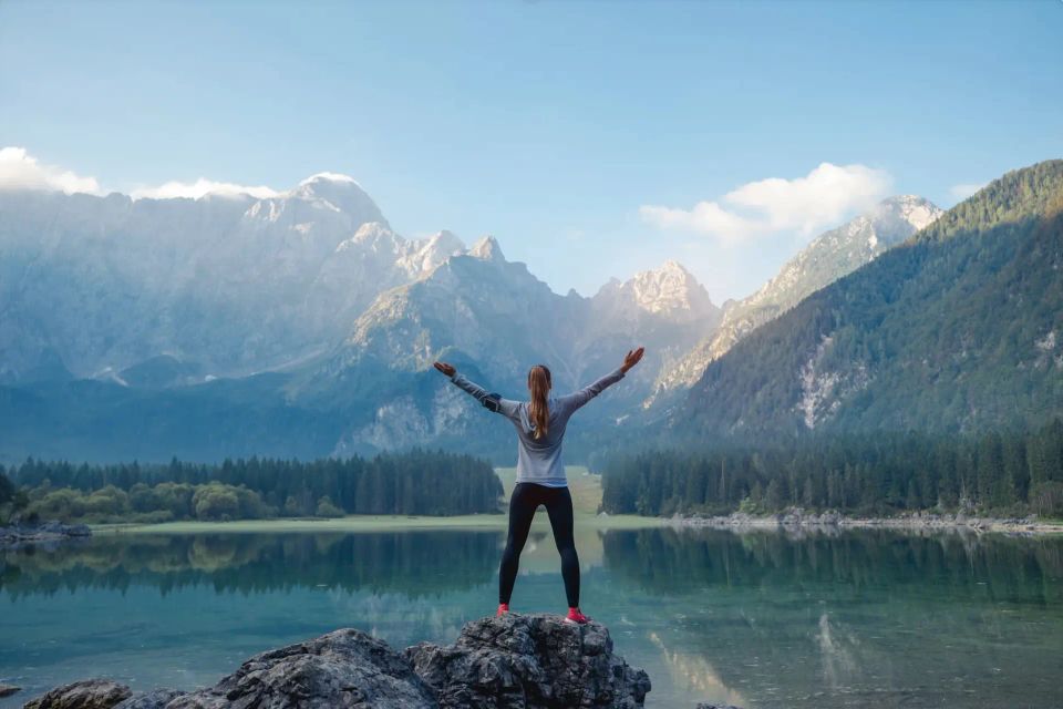 A woman stands on a rock at a mountain lake with her arms raised toward the peaks